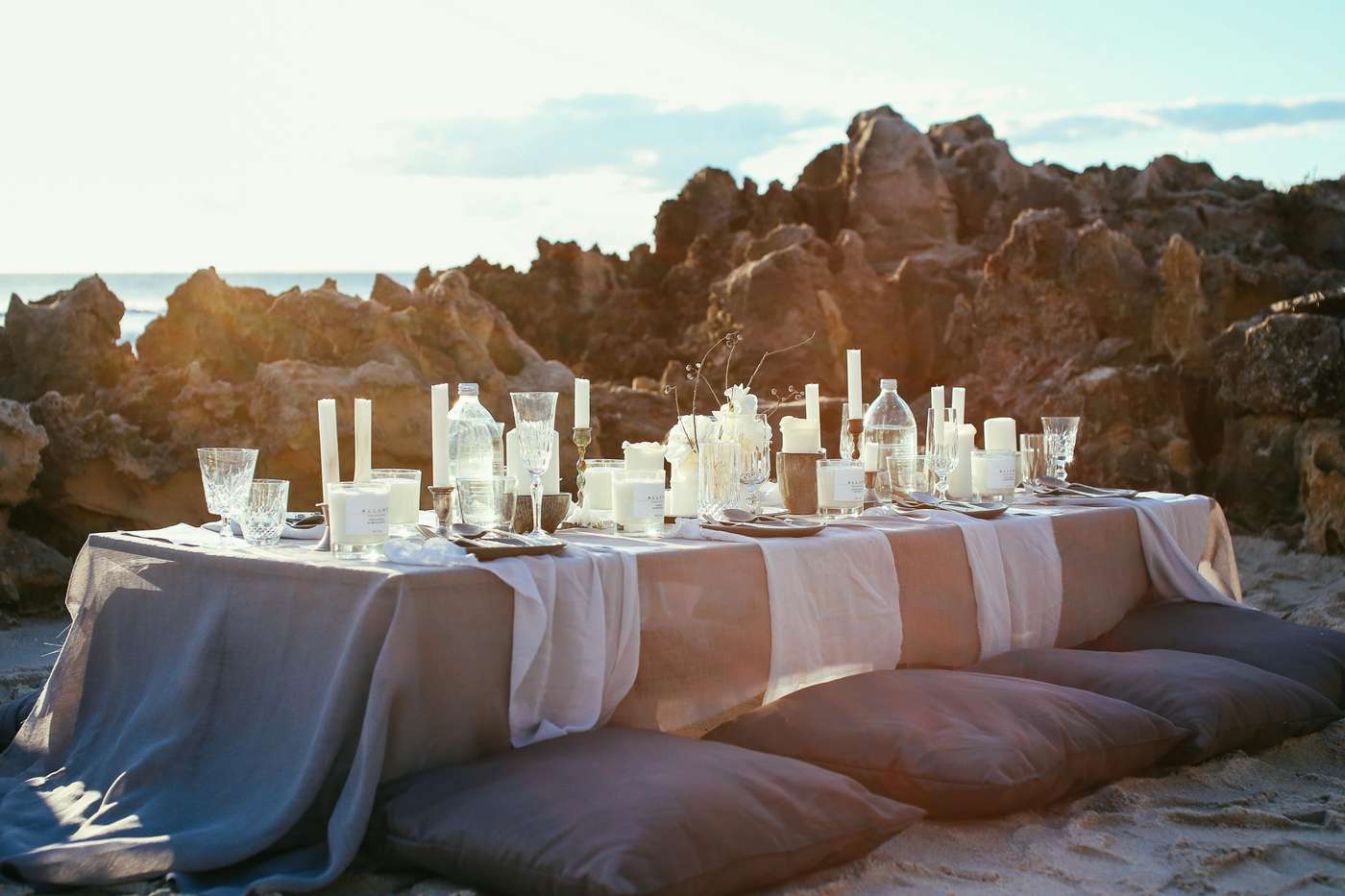 A beautifully arranged table setup on a beach with rocks and sunset in the background.