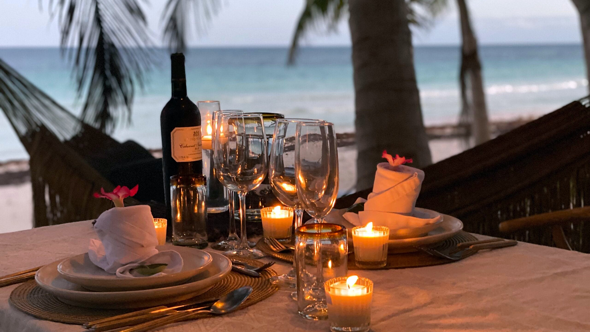 A candlelit table for two with wine glasses beside a hammock and the Caribbean at Casa Nalum